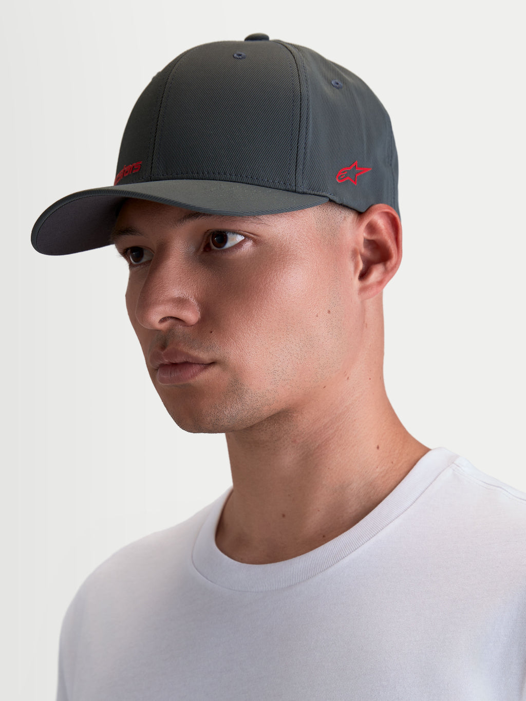 Alpinestars Casquette Points, casquette sportswear à visière incurvée portée par un modèle, gris foncé avec logos rouges, fabriquée en nylon tactel léger avec coupe flex-back pour un confort quotidien.