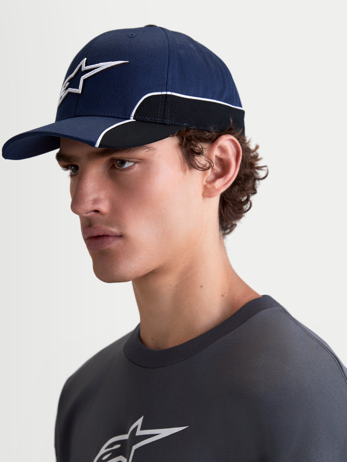 Alpinestars 500 Snapback, casquette de sportswear mixte, bleu marine et noire, vue de profil portée par un modèle. Elle possède une visière incurvée, un logo Astars brodé en 3D avec un passepoil blanc contrasté, fabriquée en sergé de coton résistant.