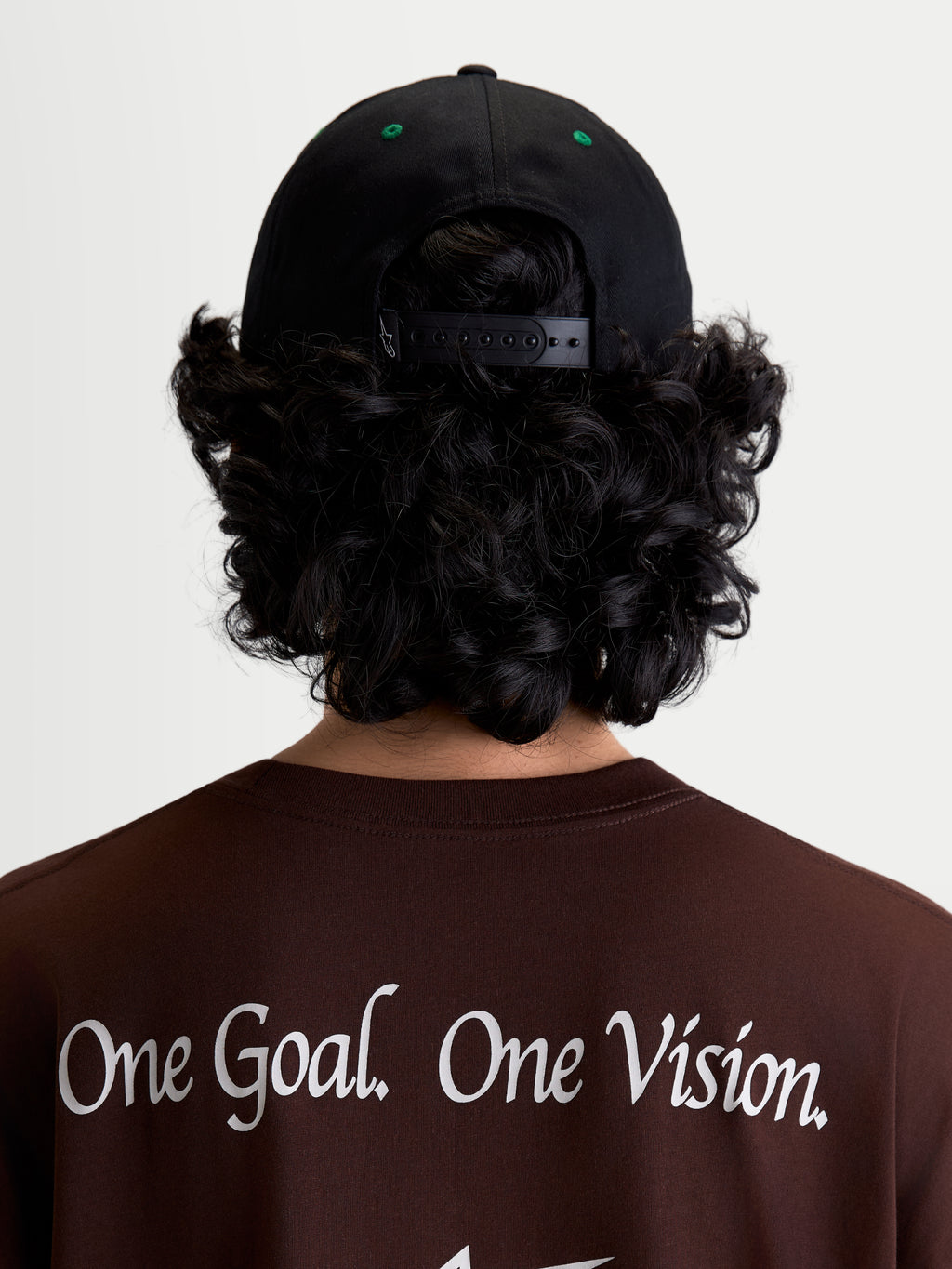 Alpinestars Casquette Retro Snapback, casquette snapback sportswear, noire et verte, vue arrière portée montrant la fermeture snapback réglable et les détails d'œillets verts, portée avec un t-shirt marron arborant le manifeste Alpinestars One Goal. One Vision.