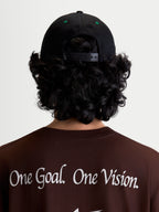 Alpinestars Casquette Retro Snapback, casquette snapback sportswear, noire et verte, vue arrière portée montrant la fermeture snapback réglable et les détails d'œillets verts, portée avec un t-shirt marron arborant le manifeste Alpinestars One Goal. One Vision.