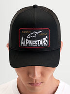 Alpinestars Casquette Trucker Hemmings, casquette snapback sportswear noire portée par un modèle, avec écusson tissé du logo sur le devant et panneaux arrière en mesh respirant