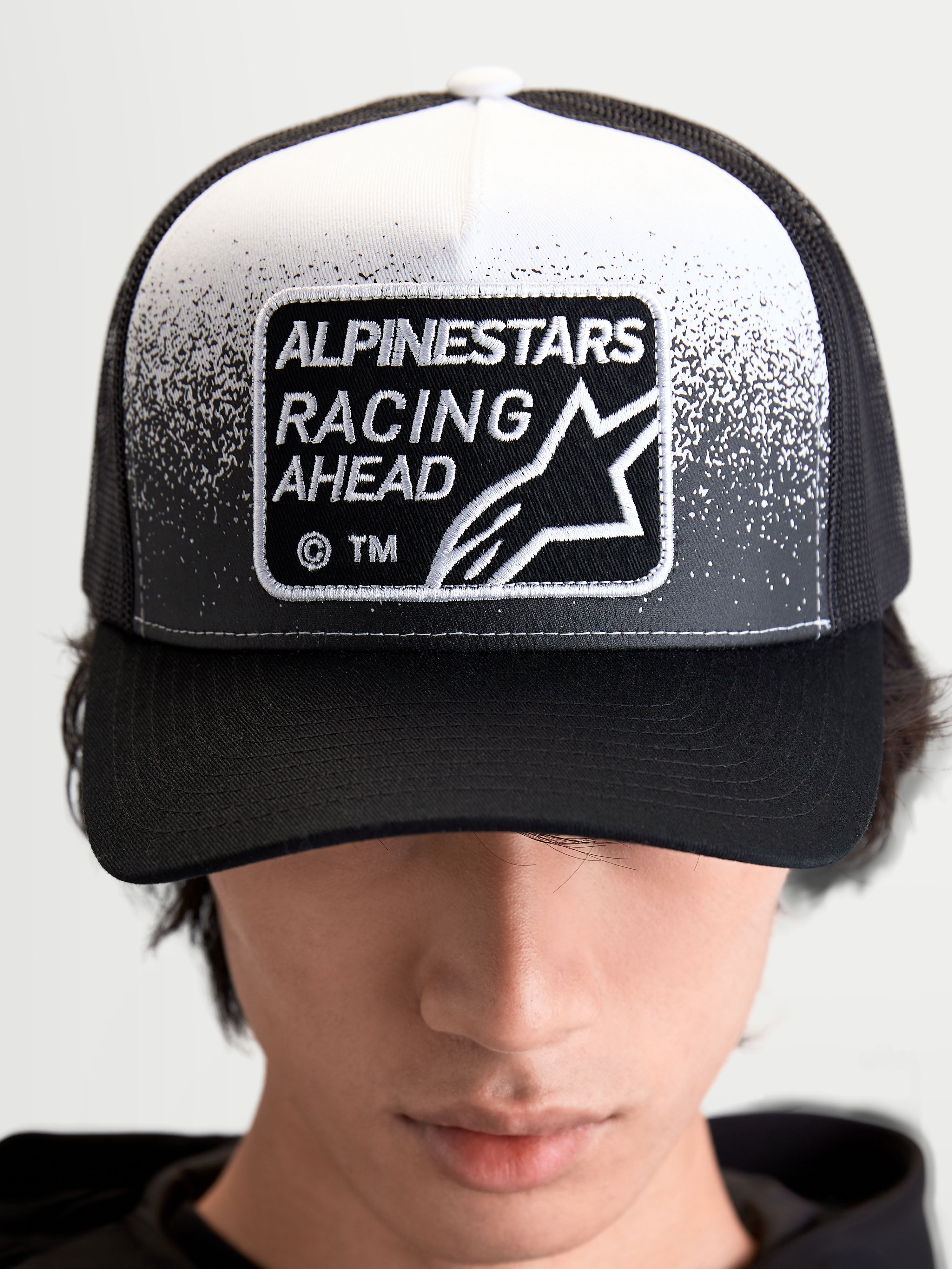 Alpinestars Casquette Trucker Gravel, casquette sportswear unisexe, noire et blanche, avec écusson tissé Alpinestars Racing Ahead à texture gravier, couronne structurée et visière courbée, vue de face sur modèle