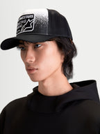 Alpinestars Casquette Trucker Gravel, casquette sportswear noire et blanche sur modèle, dotée d'une couronne structurée, d'une visière courbée, d'un arrière en maille respirante et d'un écusson tissé Alpinestars Racing Ahead à texture gravier.