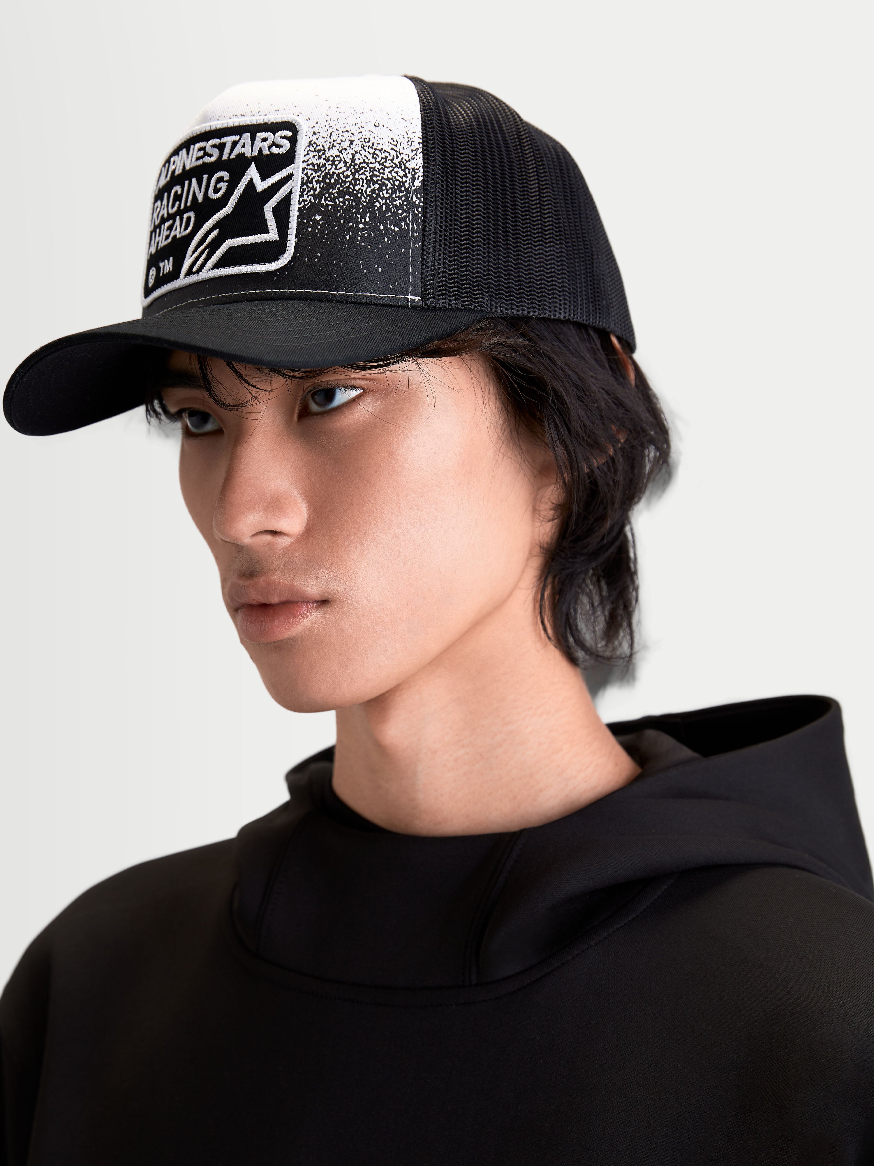 Alpinestars Casquette Trucker Gravel, casquette sportswear noire et blanche sur modèle, dotée d'une couronne structurée, d'une visière courbée, d'un arrière en maille respirante et d'un écusson tissé Alpinestars Racing Ahead à texture gravier.