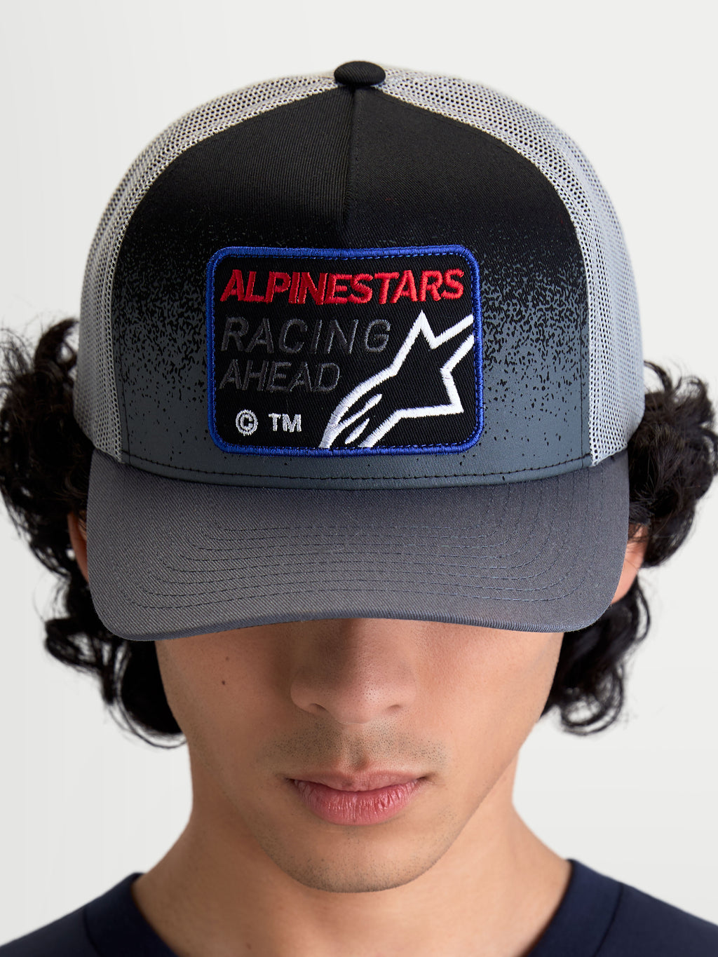 Alpinestars Casquette Trucker Gravel, casquette de sportswear, gris foncé et noir, dotée d'une visière courbée et d'un écusson frontal tissé avec le logo Astars et le texte 'Racing Ahead', portée par un modèle en vue de face.