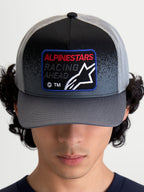 Alpinestars Casquette Trucker Gravel, casquette de sportswear, gris foncé et noir, dotée d'une visière courbée et d'un écusson frontal tissé avec le logo Astars et le texte 'Racing Ahead', portée par un modèle en vue de face.