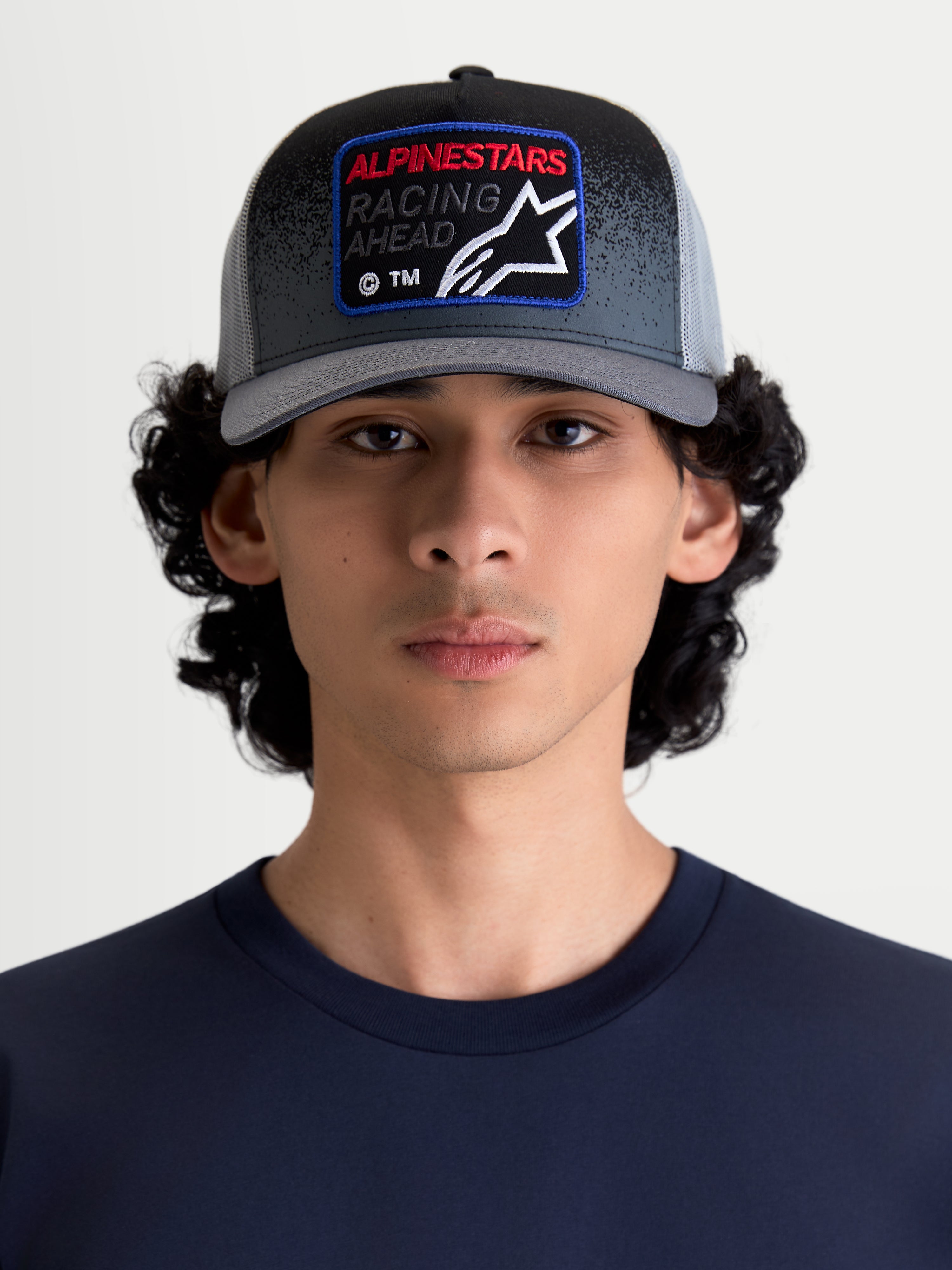 Alpinestars casquette Trucker Gravel, casquette de sportswear décontractée avec arrière en maille pour homme et femme, gris foncé et noire, portée par un modèle. Elle possède une couronne structurée, une visière courbée et un écusson tissé à l'avant avec le logo Alpinestars Racing Ahead et des détails à texture gravier.