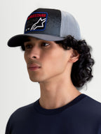 Alpinestars Casquette Trucker Gravel, casquette trucker sportswear portée par un modèle, gris foncé et noire avec arrière en maille gris clair, visière courbée et écusson tissé avec texte Alpinestars rouge, style inspiré des sports mécaniques.