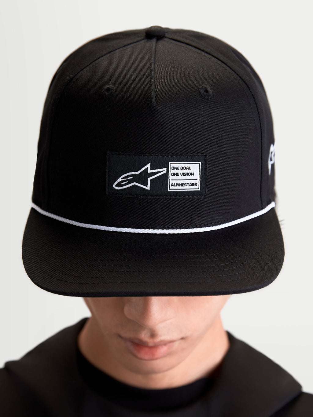 Alpinestars Label Snapback, casquette de sportswear lifestyle unisexe, noire et blanche, en sergé de coton, avec couronne structurée, visière plate, cordon blanc et écusson tissé Alpinestars, portée par un modèle
