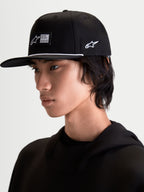 Alpinestars Casquette Label Snapback, casquette de sportswear noire et blanche avec visière plate et couronne structurée, présentant une étiquette tissée, une cordelette blanche et une broderie latérale, portée par un mannequin pour un style lifestyle moderne.