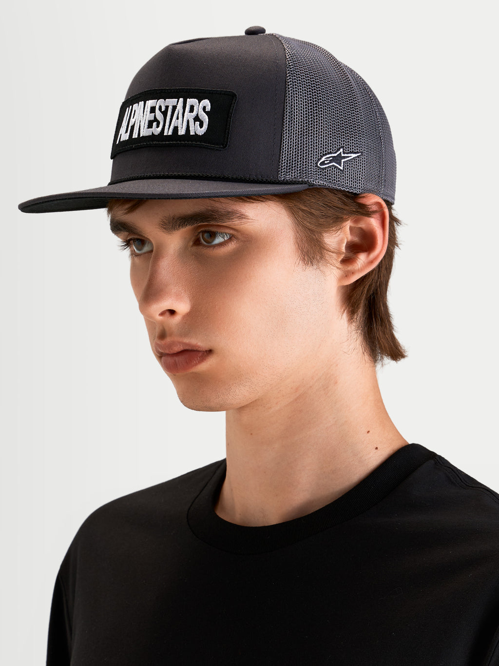Alpinestars Ten Trucker Hat, flat bill sportswear cap in dark grey, worn by a model, featuring a front embroidered Alpinestars word logo patch and side Astars logo embroidery.