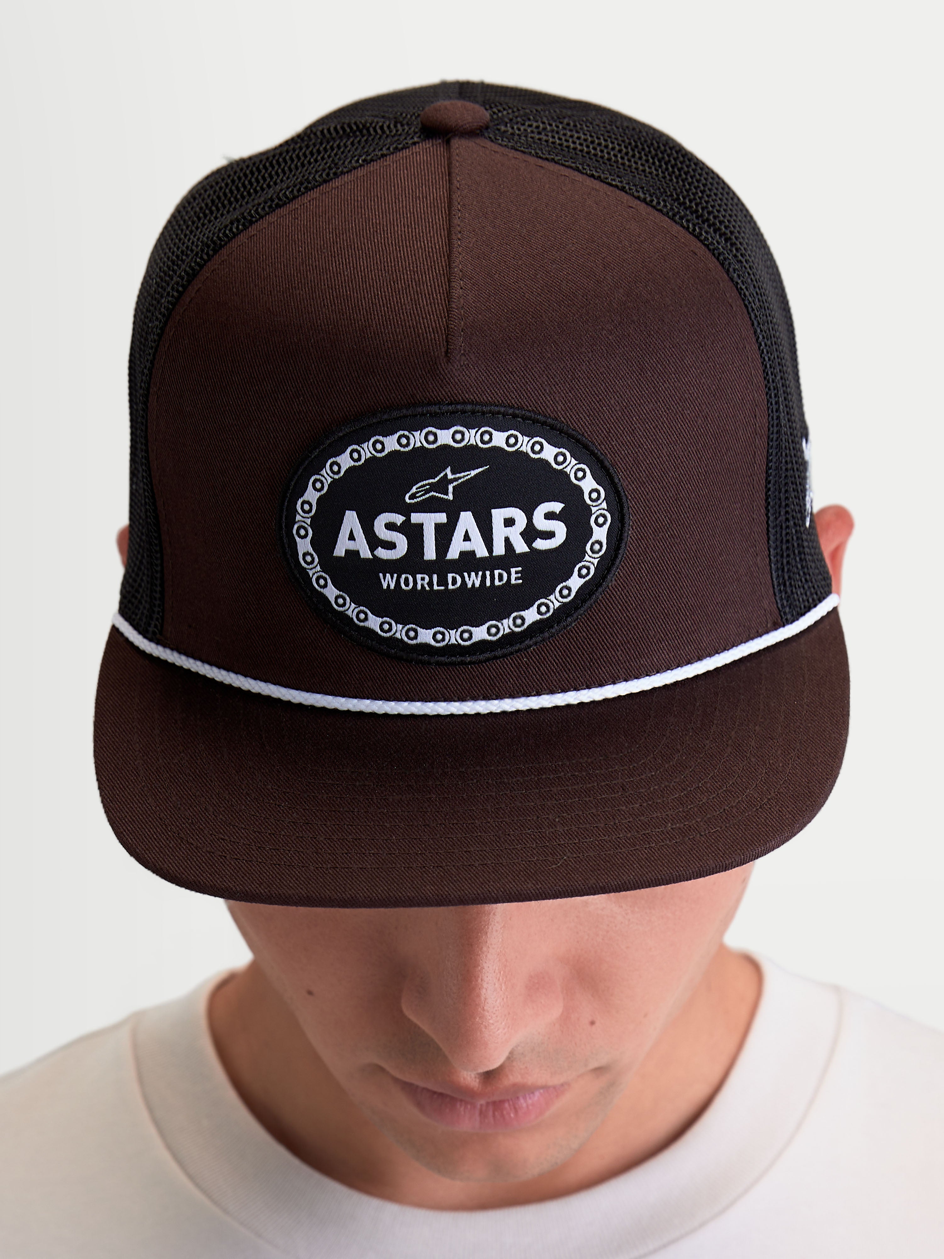 Alpinestars Casquette Trucker Hotlink, casquette de style sportswear portée par un modèle, marron et noire, couronne structurée, visière plate avec détail de cordon blanc, écusson Astars Worldwide avec bordure en chaîne de vélo.