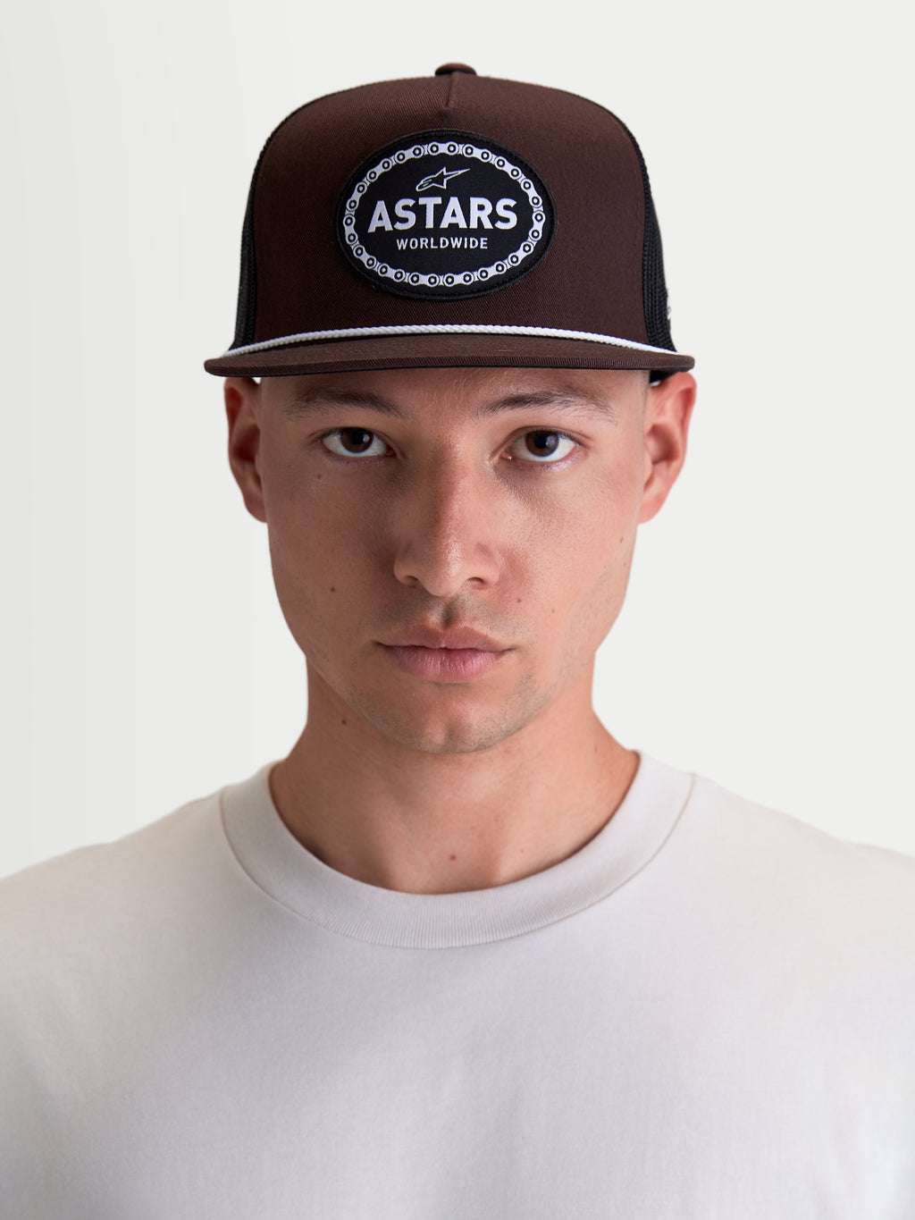 Alpinestars Casquette Trucker Hotlink, casquette sportswear inspirée du sport automobile, marron et noire, couronne structurée, visière plate avec détail de cordelette blanche, écusson Astars Worldwide à l'avant, portée par un mannequin