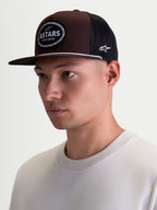 Alpinestars casquette trucker Hotlink, casquette sportswear marron et noire portée sur modèle, visière plate et couronne structurée en sergé, écusson Astars tissé avec bordure en chaîne de vélo