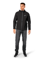 Veste de pluie Alpinestars Kascade, veste imperméable pour homme, noire, portée avec un jean gris foncé et des baskets noires, dotée d'une capuche, d'une fermeture éclair avant enduite et d'un logo Alpinestars blanc sur la poitrine, streetwear décontracté d'inspiration sport mécanique