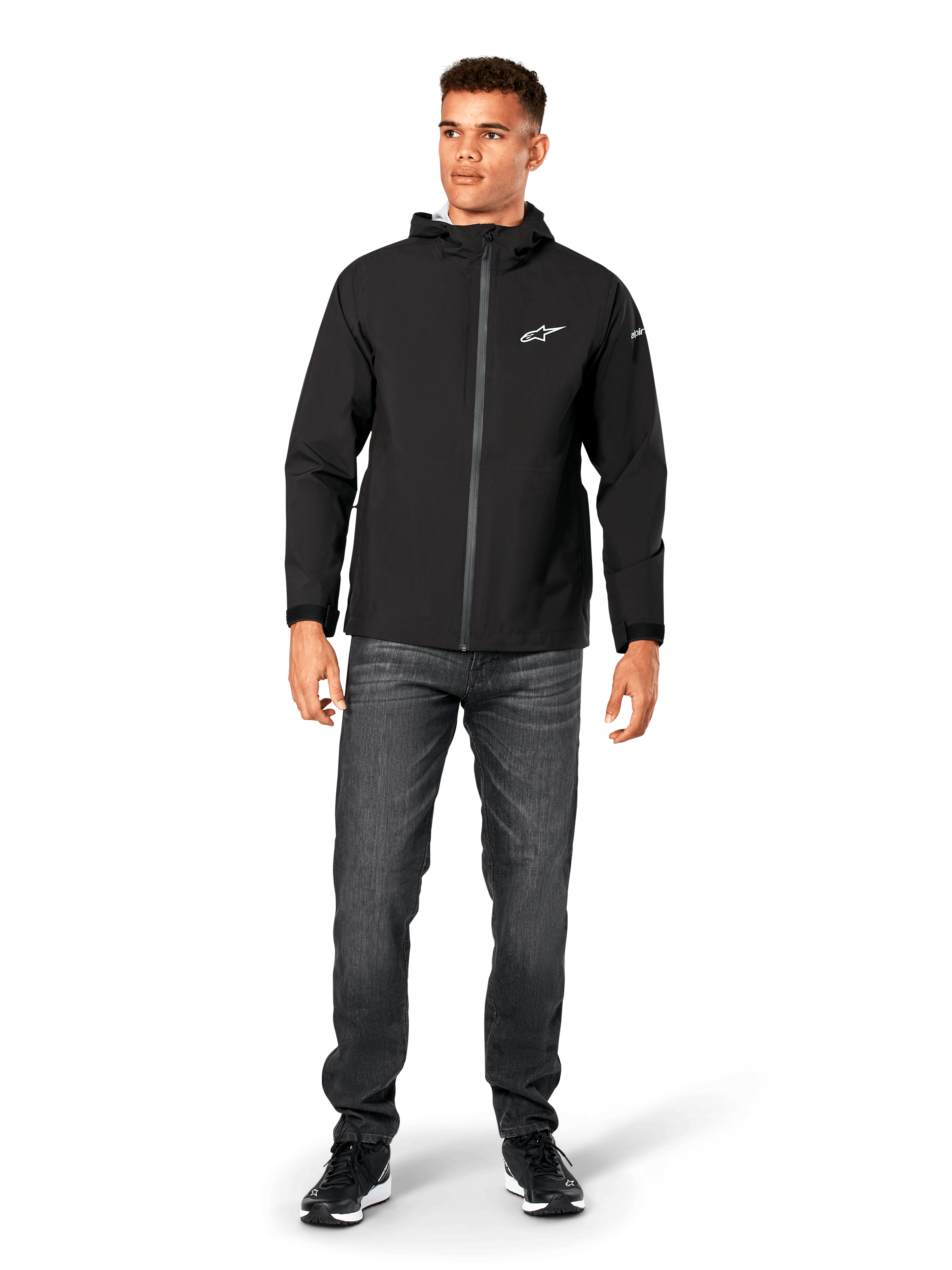 Veste de pluie Alpinestars Kascade, veste imperméable pour homme, noire, portée avec un jean gris foncé et des baskets noires, dotée d'une capuche, d'une fermeture éclair avant enduite et d'un logo Alpinestars blanc sur la poitrine, streetwear décontracté d'inspiration sport mécanique