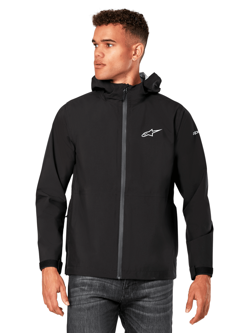 Mannequin homme portant la veste de pluie Kascade Alpinestars, noire, logos blancs sur la poitrine et la manche, veste de pluie à capuche avec fermeture éclair avant enduite et poignets Velcro, associée à un jean foncé, style streetwear décontracté inspiré des sports mécaniques