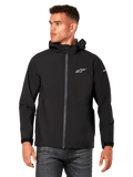 Mannequin homme portant la veste de pluie Kascade Alpinestars, noire, logos blancs sur la poitrine et la manche, veste de pluie à capuche avec fermeture éclair avant enduite et poignets Velcro, associée à un jean foncé, style streetwear décontracté inspiré des sports mécaniques