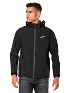 Mannequin homme portant la veste de pluie Kascade Alpinestars, noire, logos blancs sur la poitrine et la manche, veste de pluie à capuche avec fermeture éclair avant enduite et poignets Velcro, associée à un jean foncé, style streetwear décontracté inspiré des sports mécaniques