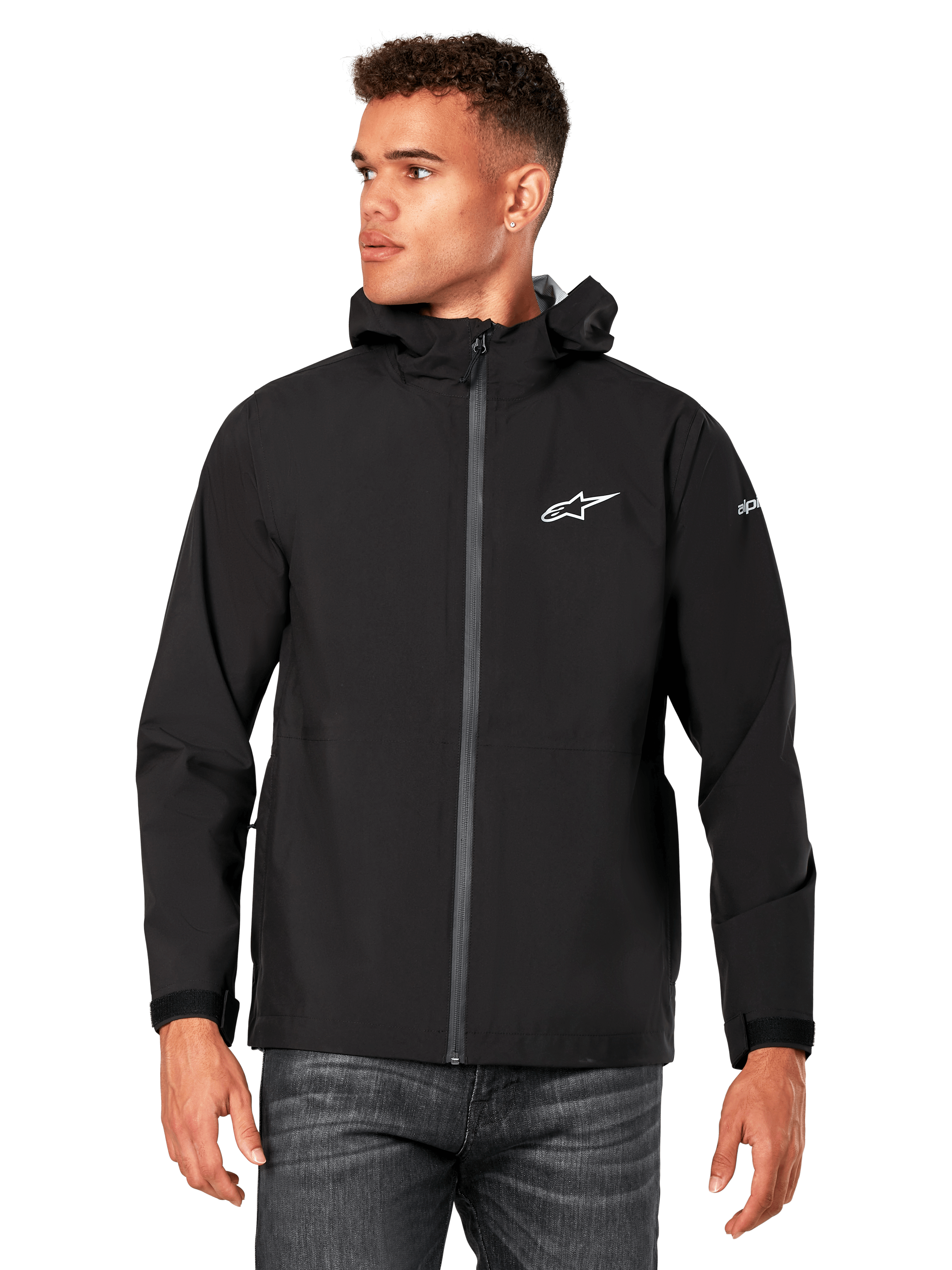 Male model wearing Alpinestars Kascade Rain Jacket, Black, white chest logo and sleeve branding, hooded rain shell with coated front zipper and velcro cuffs, paired with dark jeans, casual motorsport-inspired streetwear