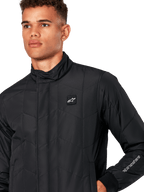 Veste Alpinestars Precedent, veste sportswear matelassée pour homme en noir, gros plan montrant le col bas, l'écusson du logo sur la poitrine et la broderie sur la manche, avec un motif matelassé en angle pour un look inspiré du sport automobile.