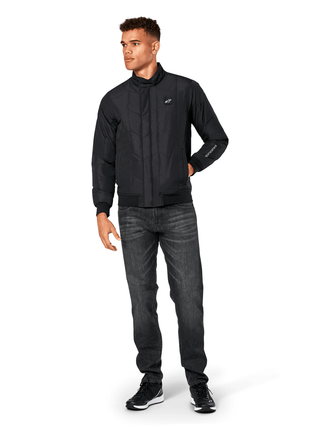 Veste Alpinestars Precedent, veste sportswear matelassée, noire, portée avec un jean gris foncé et des baskets noires, arborant un motif matelassé en angle, un col bas, des poignets et un ourlet en bords-côtes, avec un écusson logo blanc sur la poitrine et une broderie sur la manche.