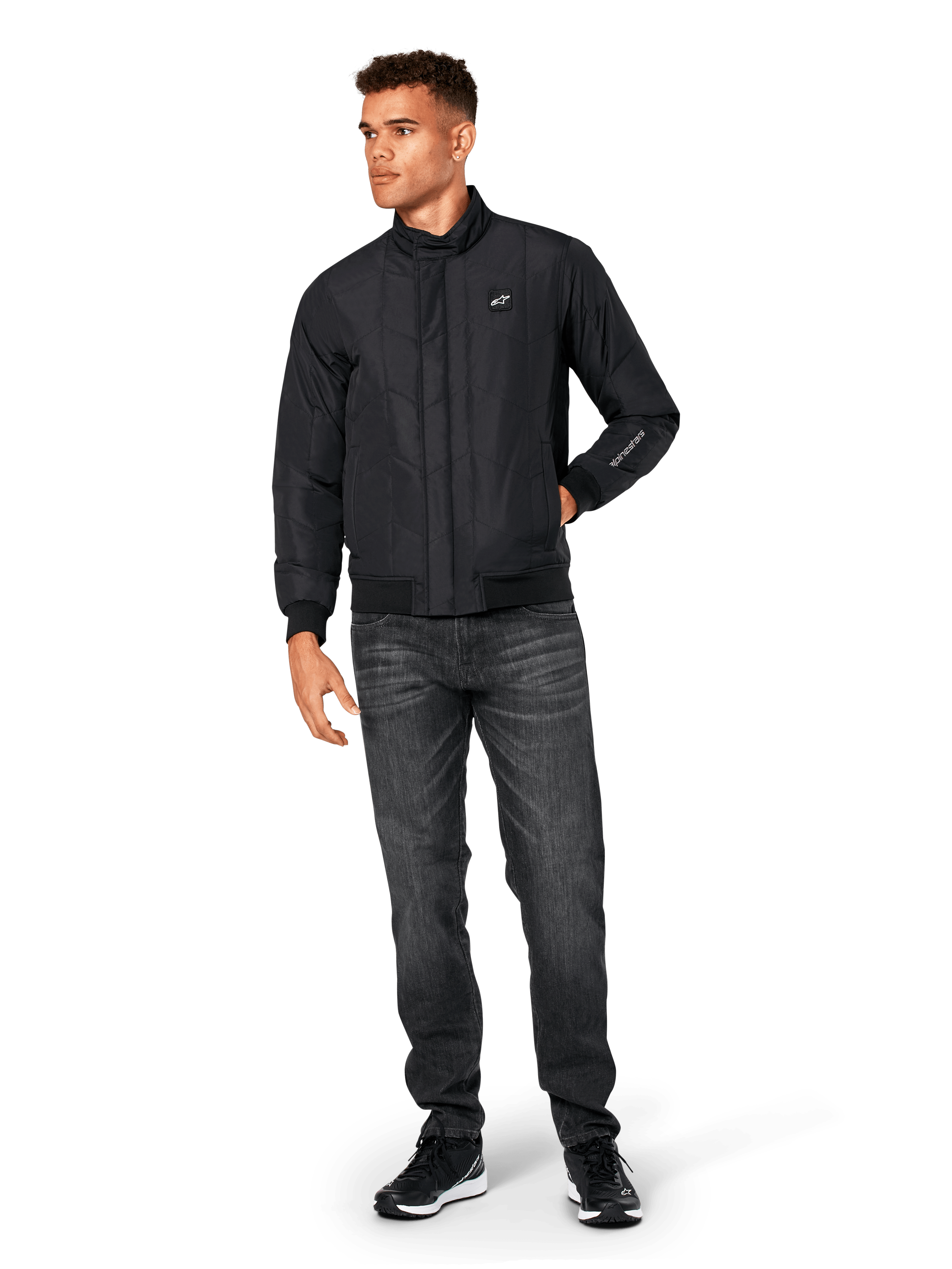 Veste Alpinestars Precedent, veste sportswear matelassée, noire, portée avec un jean gris foncé et des baskets noires, arborant un motif matelassé en angle, un col bas, des poignets et un ourlet en bords-côtes, avec un écusson logo blanc sur la poitrine et une broderie sur la manche.