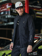 Veste Alpinestars Precedent, veste matelassée noire au style Formula 1, avec un motif matelassé en angle iconique, un écusson logo sur la poitrine et une broderie blanche sur la manche, portée avec une casquette Alpinestars assortie dans un garage de sports mécaniques.