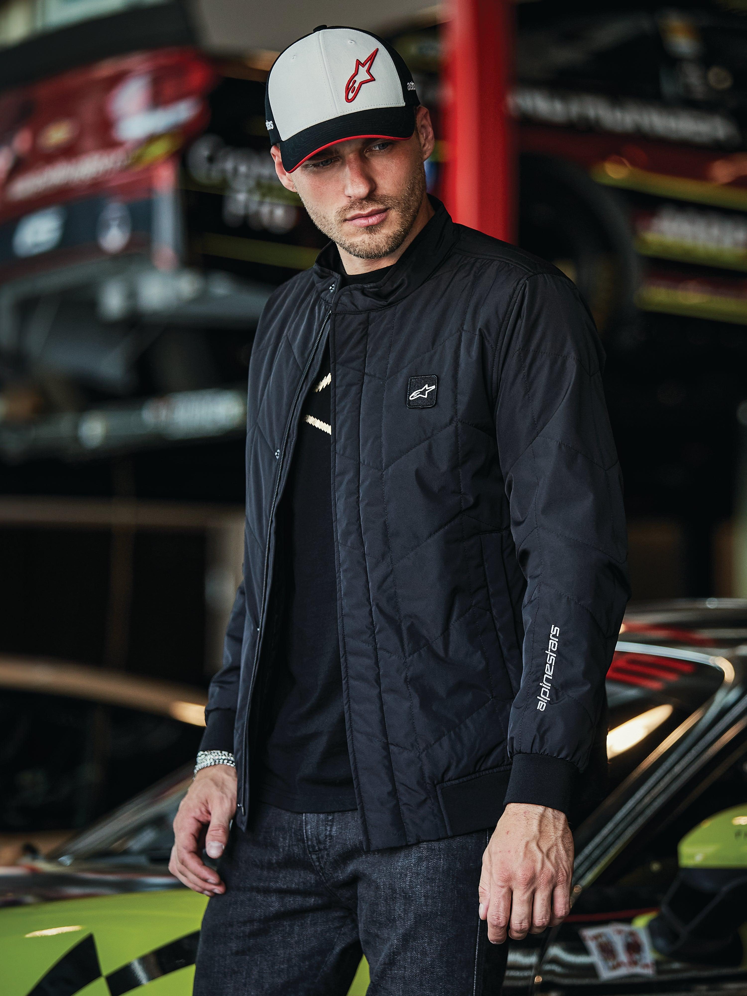 Veste Alpinestars Precedent, veste matelassée noire au style Formula 1, avec un motif matelassé en angle iconique, un écusson logo sur la poitrine et une broderie blanche sur la manche, portée avec une casquette Alpinestars assortie dans un garage de sports mécaniques.