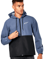 Modèle masculin portant la veste coupe-vent Alpinestars Speeder, design bicolore bleu ardoise et noir, capuche avec cordons de serrage noirs, logo blanc sur la poitrine, coupe-vent décontracté d'inspiration sport mécanique