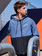 Veste coupe-vent Alpinestars Speeder, coupe-vent léger à capuche, design bicolore bleu ardoise et noir, logo Alpinestars blanc sur la poitrine, doublure en mesh et capuche à cordon de serrage, streetwear décontracté inspiré du sport automobile