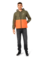 Veste coupe-vent Alpinestars Speeder, bicolore vert militaire et orange, veste à capuche légère avec cordons de serrage noirs et fermeture éclair avant, logo blanc sur la poitrine et la manche, portée avec un jean foncé et des baskets noires, style sportswear décontracté