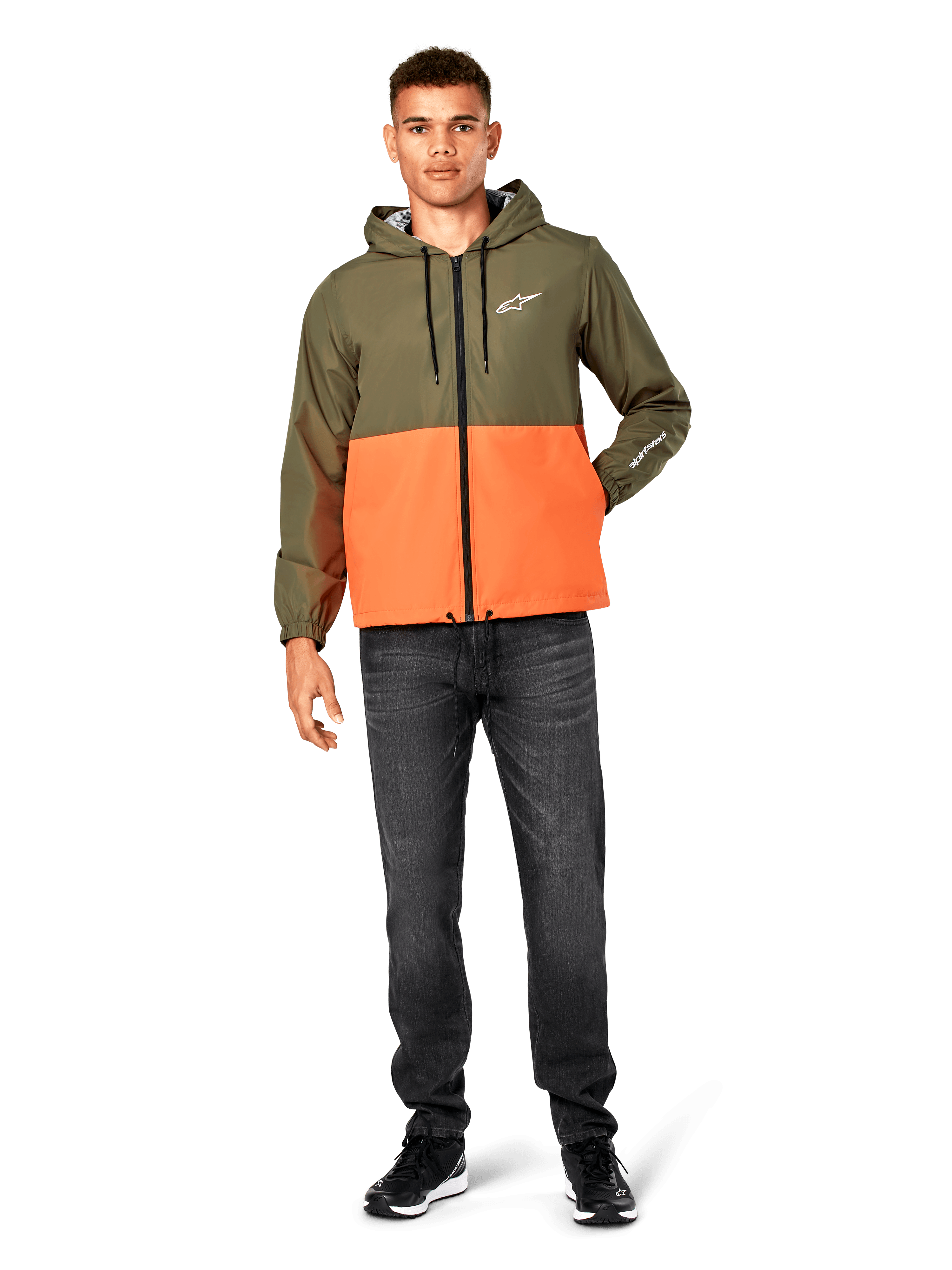 Veste coupe-vent Alpinestars Speeder, bicolore vert militaire et orange, veste à capuche légère avec cordons de serrage noirs et fermeture éclair avant, logo blanc sur la poitrine et la manche, portée avec un jean foncé et des baskets noires, style sportswear décontracté