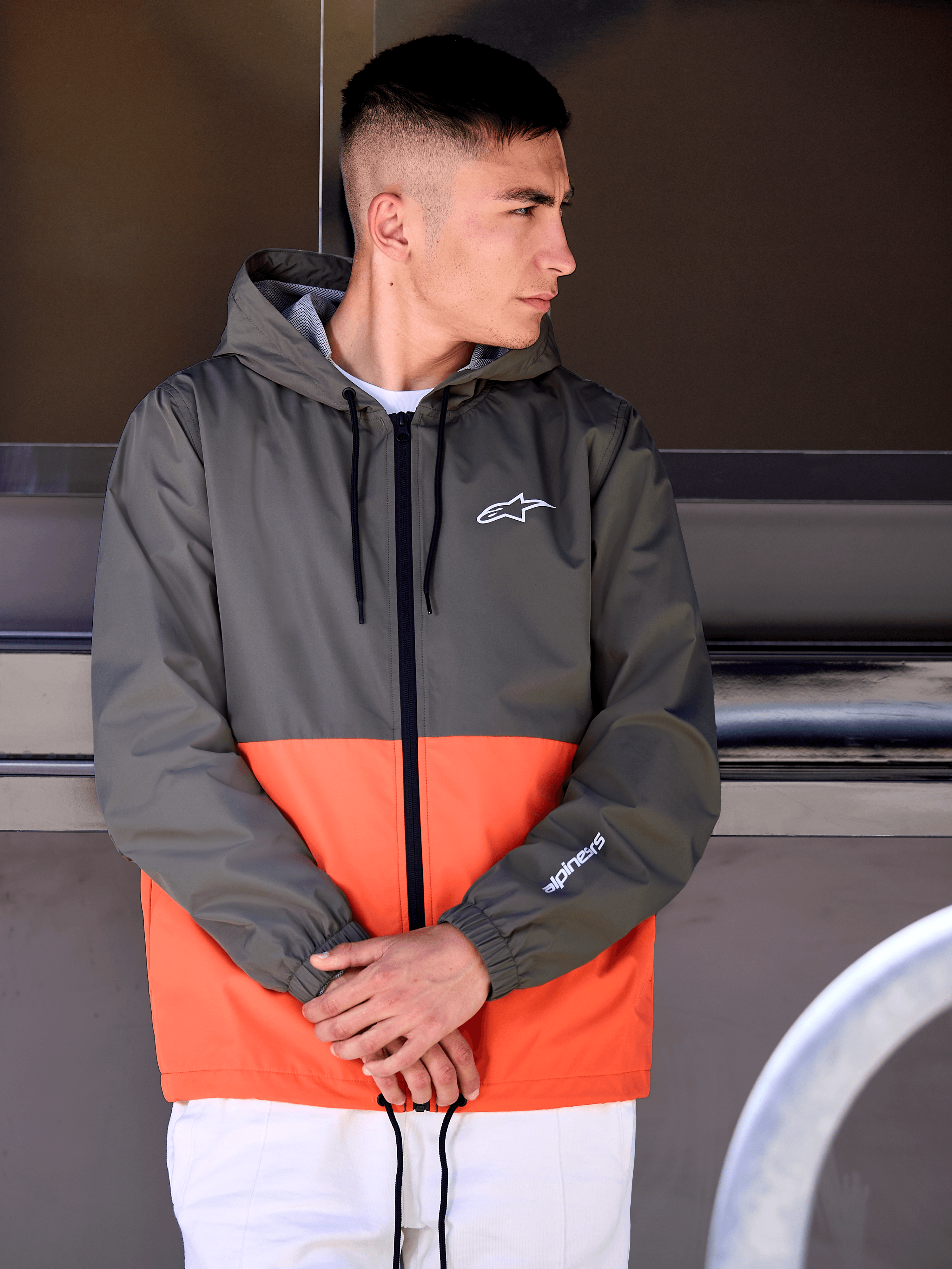 Homme portant la veste coupe-vent Alpinestars Speeder, design color-block orange militaire et vert foncé, vêtement d'extérieur décontracté à capuche avec logos blancs sur la poitrine et la manche, associé à un pantalon blanc pour un look lifestyle inspiré du sport automobile