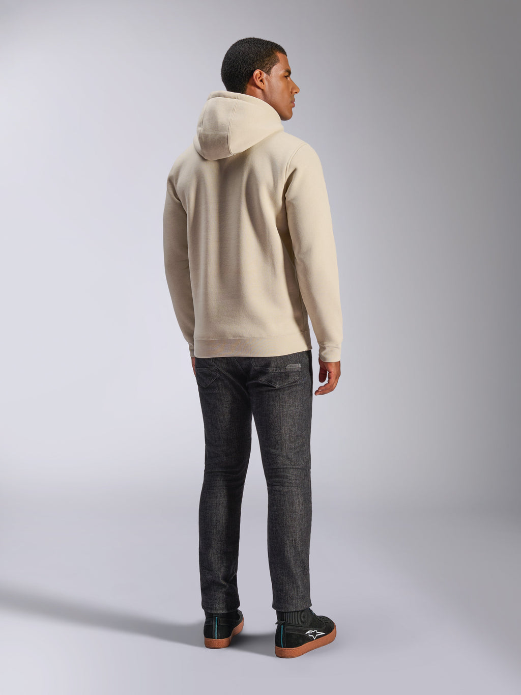 Sweat à capuche Alpinestars Ageless V3, sweat décontracté en polaire blanc cassé vu de dos, avec poignets et ourlet côtelés, porté avec un jean slim foncé et des baskets Alpinestars noires