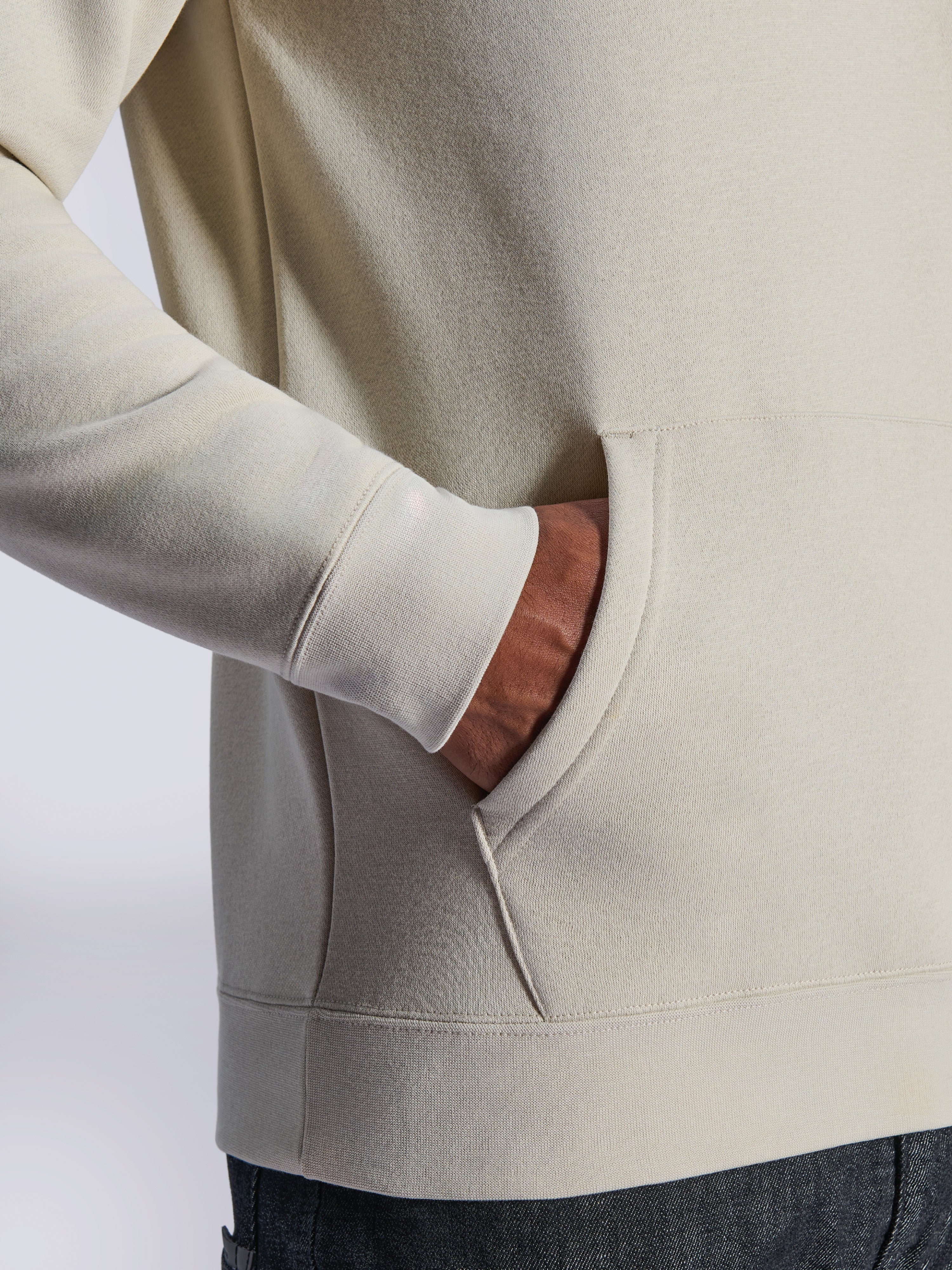 Sweat à capuche Alpinestars Ageless V3, détail en gros plan de la poche kangourou avant et du poignet côtelé, polaire blanc cassé, streetwear décontracté d'inspiration motorsport
