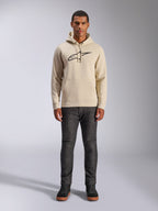 Sweat à capuche Alpinestars Ageless V3, vêtement sportswear décontracté, blanc cassé avec logo brodé noir, porté par un homme avec un pantalon gris foncé et des baskets noires, doté d'une capuche avec cordon de serrage et d'une poche kangourou, design moderne inspiré par le sport automobile