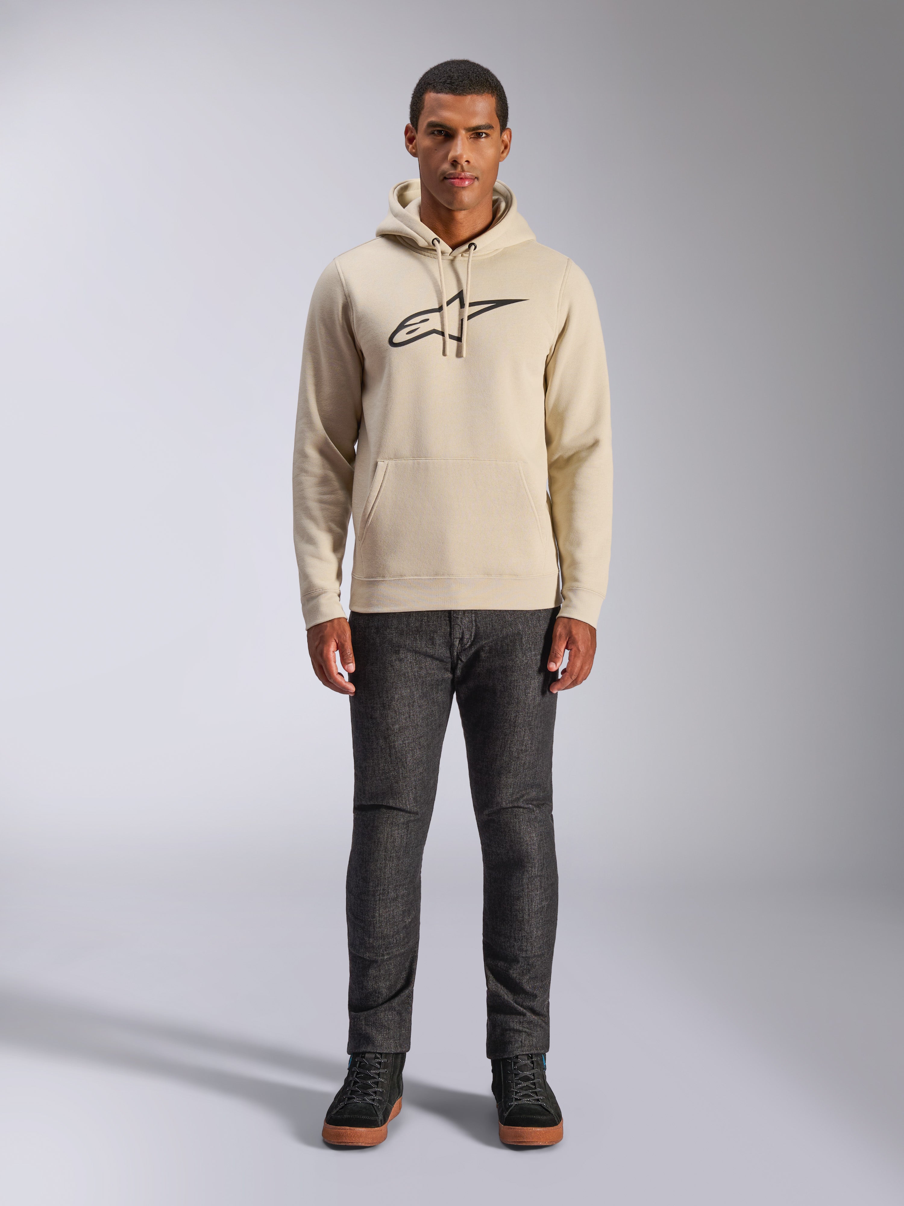 Sweat à capuche Alpinestars Ageless V3, vêtement sportswear décontracté, blanc cassé avec logo brodé noir, porté par un homme avec un pantalon gris foncé et des baskets noires, doté d'une capuche avec cordon de serrage et d'une poche kangourou, design moderne inspiré par le sport automobile