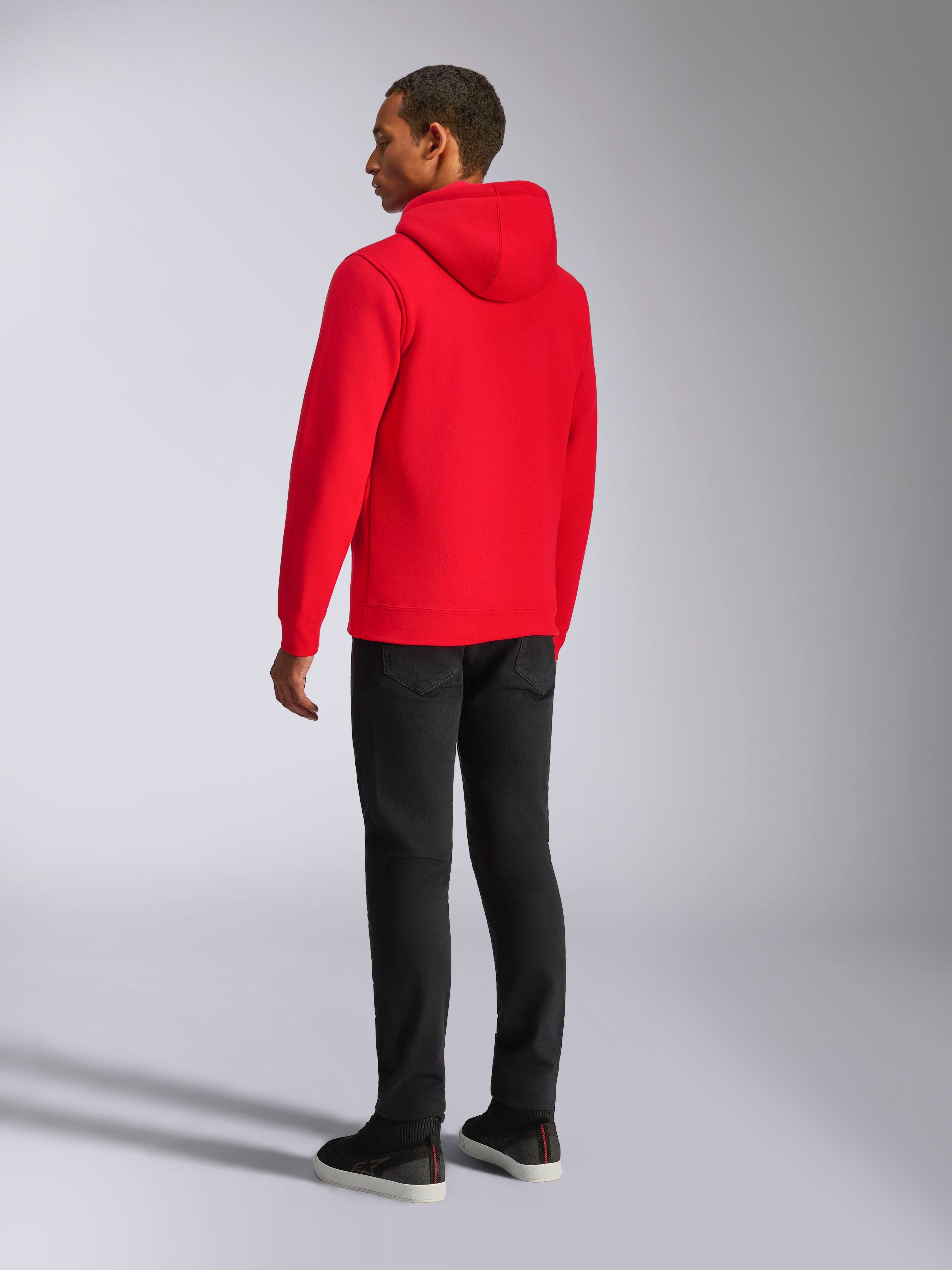 Sweat à capuche Alpinestars Ageless V3, streetwear décontracté, rouge vif, modèle de dos, avec capuche doublée en jersey, poignets et ourlet côtelés, porté avec un pantalon noir, design moderne inspiré du sport automobile