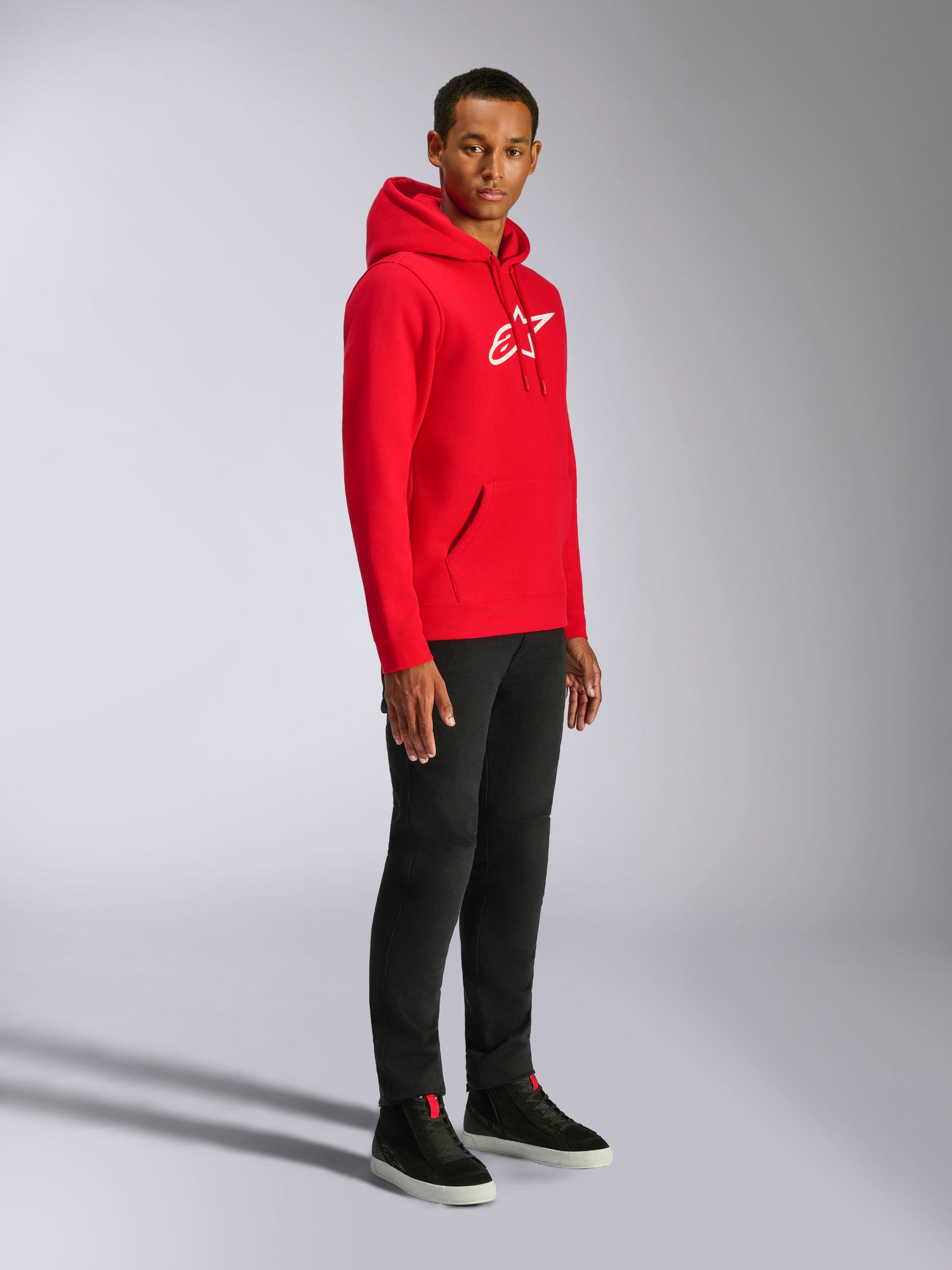 Sweat à capuche Alpinestars Ageless V3, rouge avec logo blanc brodé, sweat sportswear décontracté porté par un homme avec un pantalon noir et des baskets noires, doté d'une capuche à cordon de serrage et de poignets côtelés