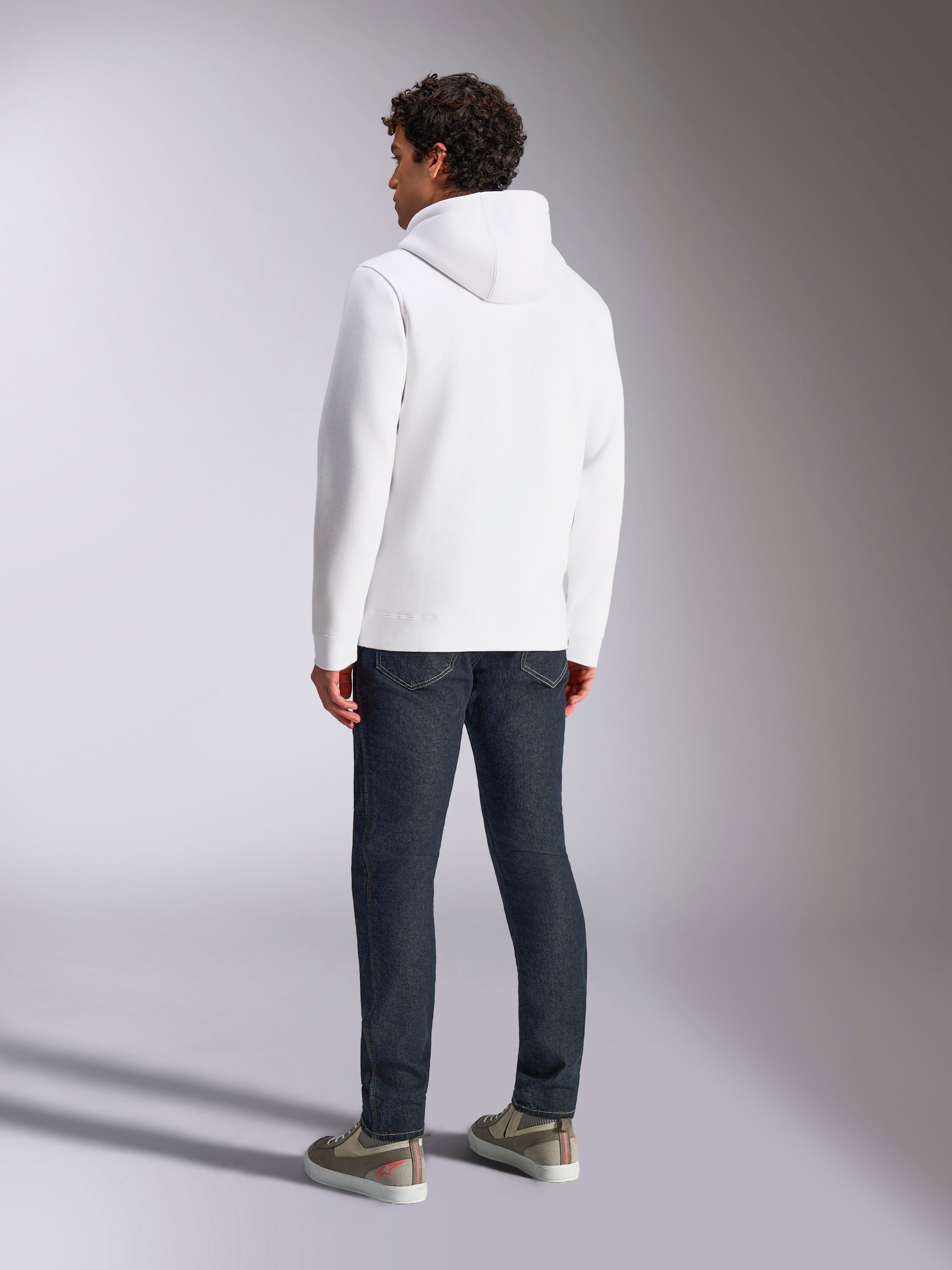 Alpinestars Blaze V3 Hoodie, vue arrière d'un mannequin portant un sweat à capuche blanc, style sportswear décontracté avec un jean bleu foncé et des baskets grises, design moderne inspiré des sports mécaniques