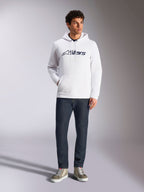 Sweat à capuche Alpinestars Blaze V3, sweat à capuche blanc de style streetwear décontracté avec logo Alpinestars bleu marine sur la poitrine, porté par un mannequin avec un jean bleu foncé et des baskets beiges, doté d'une capuche à cordon de serrage et d'une poche kangourou à l'avant