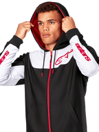 Alpinestars Sessions V3 Hoodie, noir, blanc et rouge, sweat à capuche zippé porté par un homme avec la capuche relevée, logos Alpinestars rouges sur la poitrine et les manches, streetwear décontracté de sport automobile
