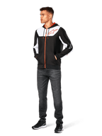 Sweat à capuche Alpinestars Sessions V3, sweat à capuche zippé décontracté, noir, blanc et rouge, porté par un mannequin avec un jean foncé et des baskets noires, doté d'empiècements blancs sur la poitrine et les manches, détails de fermeture éclair et cordons de serrage rouges, grand logo sur la poitrine, polaire en polyester au style streetwear inspiré du sport automobile