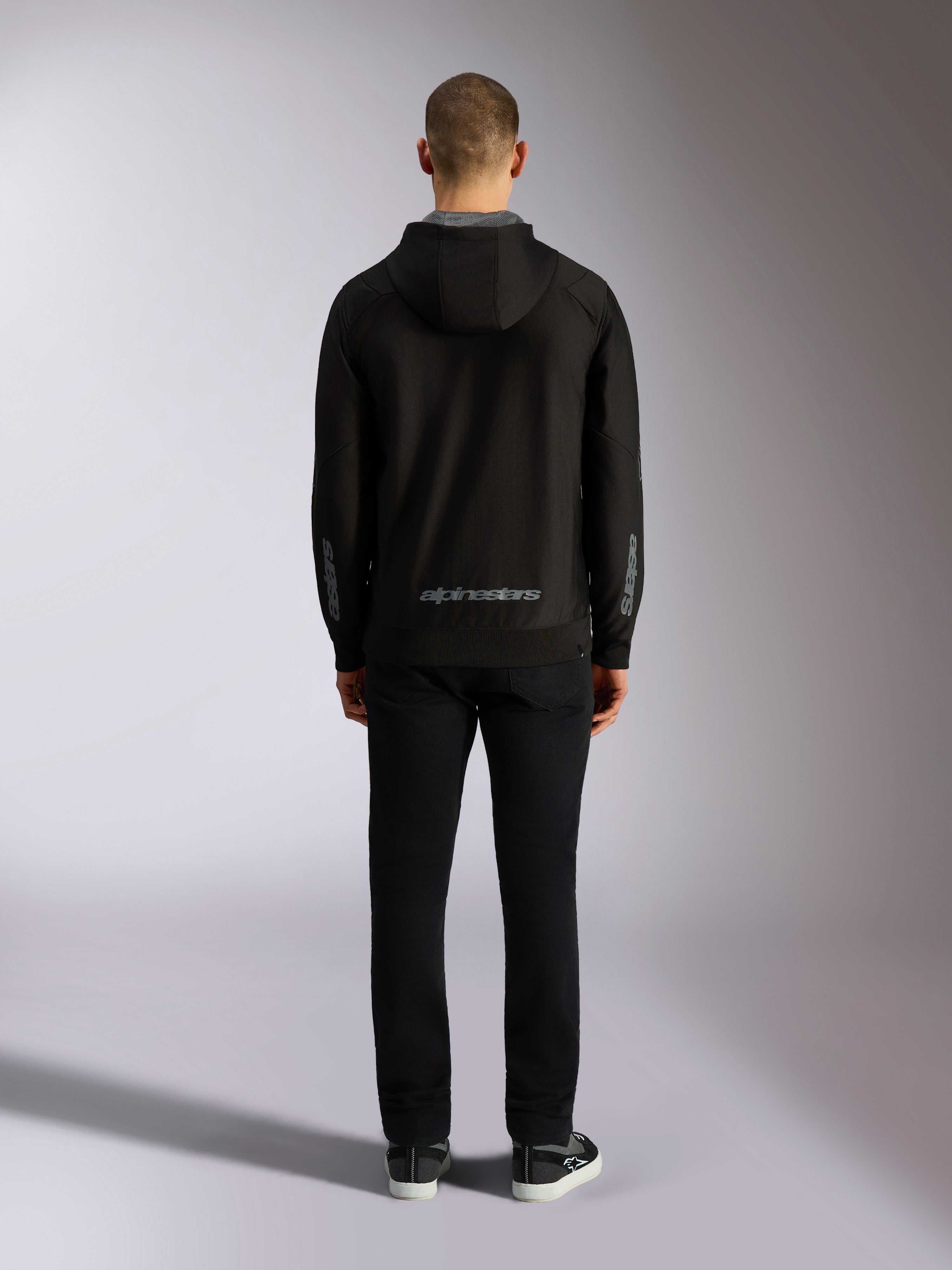 Back view of Alpinestars Sessions V3 Hoodie in Black Grey, featuring a grey Alpinestars wordmark on the lower back and astars logos on the forearms, worn by a model with black pants, casual motorsport-inspired streetwear