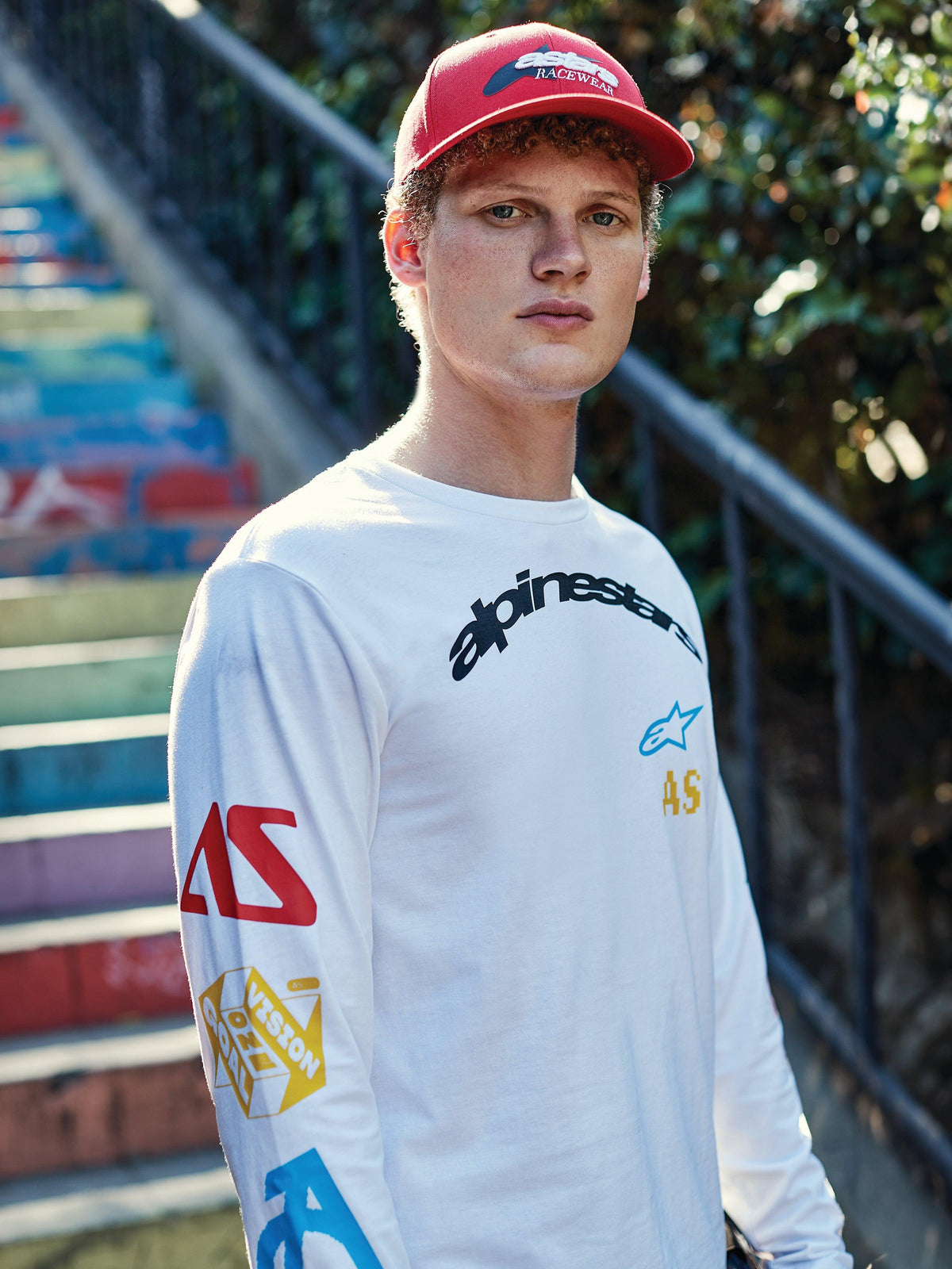 Alpinestars Brundo CSF Long Sleeve Tee, White, featuring black chest branding and multi-colored graphics on the sleeves, worn by a man in a red cap, casual motorsport streetwear style.