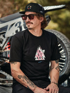 Casquette Trucker Alpinestars Gambit en noir avec écusson logo circulaire, portée avec le t-shirt Alpinestars Soloist SS noir, photo lifestyle mettant en scène un homme avec des lunettes de soleil et des tatouages assis devant une moto de motocross.