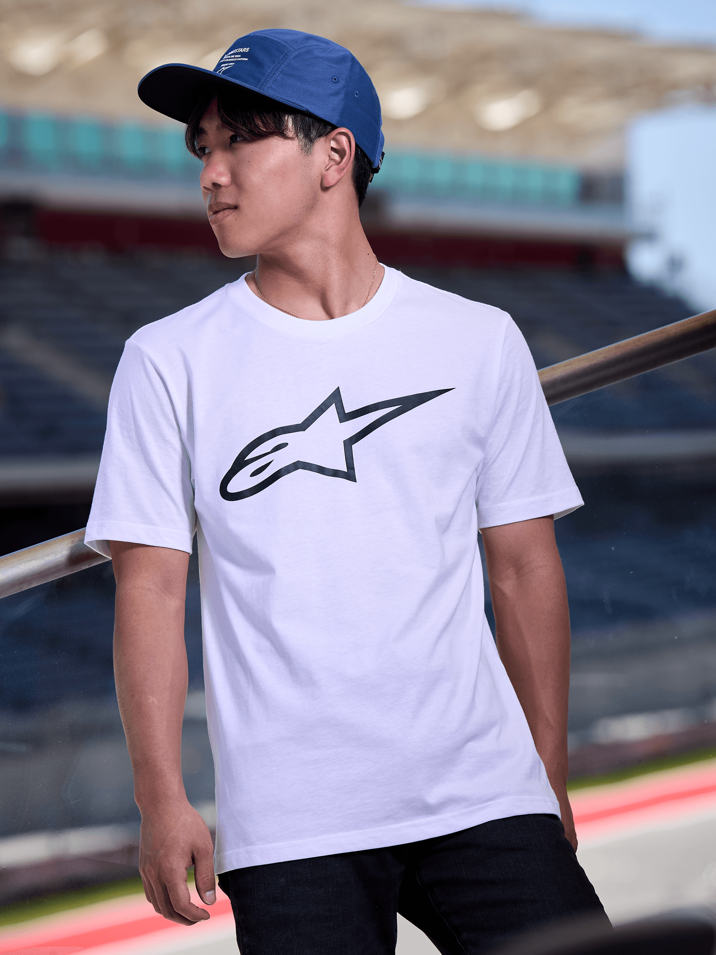Homme portant la casquette Alpinestars Peasy en bleu, casquette jockey à 5 panneaux avec visière plate et logo graphique blanc imprimé, associée à un t-shirt blanc avec logo Alpinestars, style streetwear décontracté inspiré des sports mécaniques