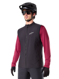 Gilet thermique Alpinestars A-Dura, noir, gilet thermique de cyclisme, porté sur un maillot à manches longues bordeaux, avec un logo blanc sur la poitrine, associé à un casque de VTT noir et des lunettes de soleil, couche de performance isolante pour le VTT