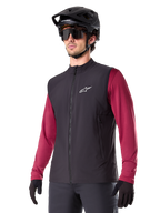 Gilet thermique Alpinestars A-Dura, noir, gilet thermique de cyclisme, porté sur un maillot à manches longues bordeaux, avec un logo blanc sur la poitrine, associé à un casque de VTT noir et des lunettes de soleil, couche de performance isolante pour le VTT
